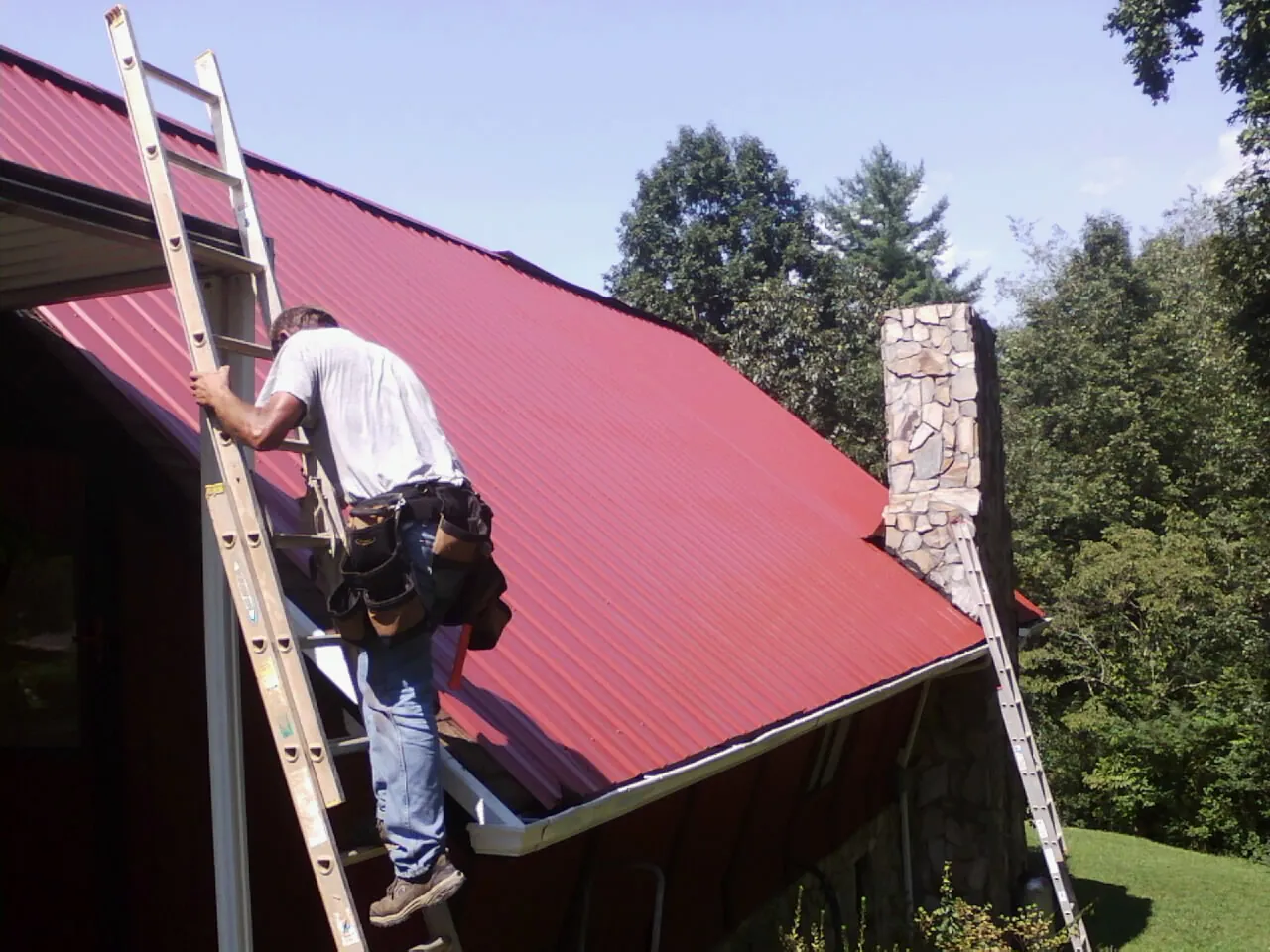 Roofer on a ladder beside a red metal roof and stone chimney — Soffit & Fascia Repair by Roofing Brothers in Glens Falls